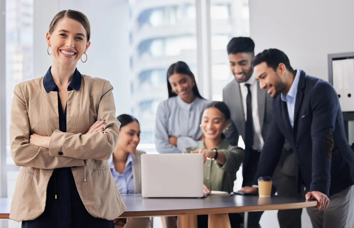 Confident hiring manager with arms crossed smiling in the foreground, while her team reviews candidates together on a laptop behind her